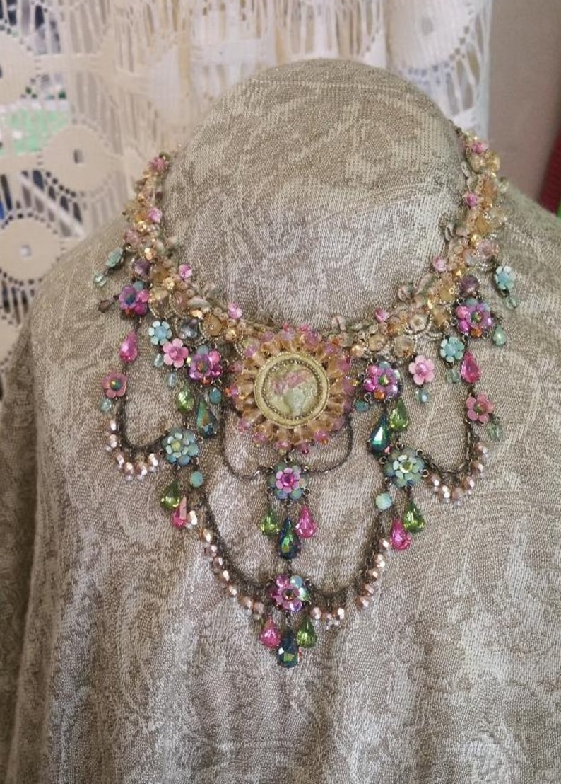 May include: A multi-layered necklace with a large, ornate centrepiece. The necklace is made of gold-toned metal and features pink, green, and blue crystals, as well as small flowers.