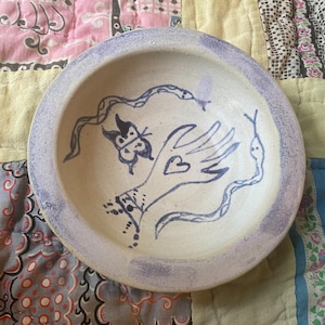 May include: A handmade ceramic bowl with a pale purple rim and a cream interior. The bowl is decorated with blue illustrations of a hand with a heart, a butterfly, and two snakes. The bowl is round and shallow.
