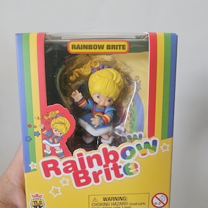 May include: A Rainbow Brite collectible figurine inside a rectangular cardboard box. The box features a yellow background with rainbow stripes, the text Rainbow Brite, and a graphic of the character. The figurine has yellow hair and a blue dress.