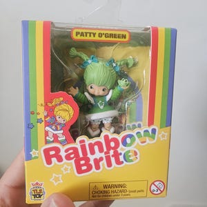 May include: A Patty O'Green Rainbow Brite collectible figure in its original packaging. The box features a yellow background with rainbow stripes, the text "Rainbow Brite" and "Patty O'Green," and a small green frog accessory inside the clear window.