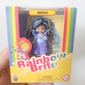May include: A Rainbow Brite Indigo character figurine in a yellow box with rainbow side panels. The doll has black hair with blue streaks, a purple outfit with a star emblem, and light blue accents. The packaging features the text Rainbow Brite.
