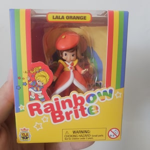 May include: A Rainbow Brite Lala Orange collectible figure inside a yellow cardboard box with rainbow stripes. The packaging features the text Rainbow Brite, Lala Orange, and a warning label about small parts and a choking hazard for children.