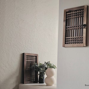 May include: Two wooden framed panels with a lattice design, one larger than the other, are mounted on a white textured wall. A white vase with a bulbous shape sits on a shelf below the smaller panel, with a small potted plant beside it.