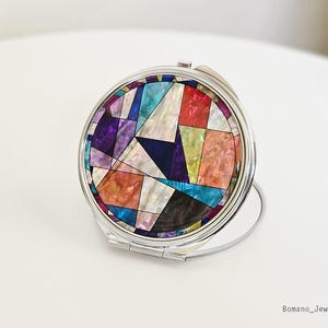 May include: A round, silver-toned compact mirror with a colorful mosaic design. The mosaic features geometric shapes in shades of purple, blue, orange, and white. The mirror is open, revealing the reflective surface. The brand name "Bomano_Jewerls" is printed on the bottom right.