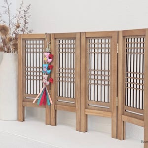 May include: A miniature wooden folding screen with a traditional Korean design. The screen is light brown with a grid pattern and white backing. A colorful ornament hangs from the screen, and a vase with dried flowers is to the left.
