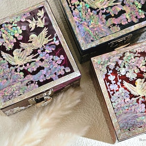 May include: Three decorative boxes with intricate floral designs and birds made from mother of pearl inlay. The boxes are in shades of black, red, and purple.