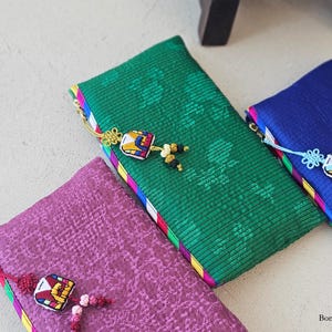 May include: Three colorful, rectangular pouches with intricate patterns and decorative tassels. The pouches are in shades of green, purple, and blue, with contrasting trim. The image is well-lit, showcasing the details of the fabric and embellishments.