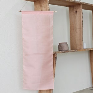 May include: A pale pink fabric banner hanging on a wooden shelf. The banner is made of a sheer, lightweight material and has a subtle sheen.