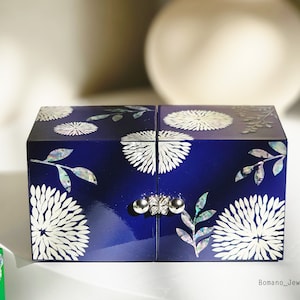 May include: A navy blue jewelry box with a glossy finish. The box is decorated with white floral and leaf designs. The box has a silver clasp and hinges. The box is a square shape and has four sections.