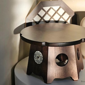 May include: A dark brown wooden side table with a round top and a decorative floral design. The table has a unique shape and a circular cutout on the side. A decorative wooden lantern is in the background.
