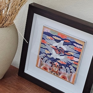May include: A framed artwork featuring a colorful embroidered design of two white cranes flying in a blue sky with clouds. The embroidery is framed in a dark brown wood frame with a white mat.