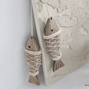May include: Two wooden fish ornaments hanging from a string with a white rope net around them. The fish are brown and have black eyes. The background is a light beige textured surface.