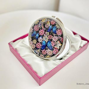 May include: A round, silver compact mirror with a colorful butterfly and floral design on a black background. The compact is open, revealing a mirror. It rests in a pink box lined with white fabric.