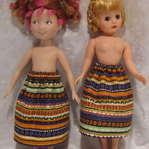 May include: Two dolls wearing colorful skirts with a Halloween theme. The skirts have orange, black, green, and purple stripes with a pattern of pumpkins and leaves.