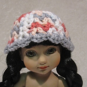 May include: A doll wearing a crocheted hat with pink, white, and gray yarn. The hat has a scalloped edge.