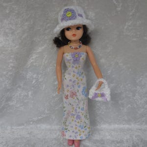May include: A doll wearing a white floral print dress, matching hat, and purse. The dress features a strapless design and a floral pattern in shades of purple, pink, and blue. The hat and purse are white with purple floral accents.