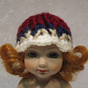 May include: A doll wearing a red, white, and blue crocheted hat. The hat has a white brim and is decorated with stripes of red, white, and blue yarn.