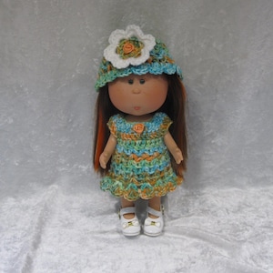 May include: A handmade doll wearing a crocheted dress and hat. The dress is a mix of blue, orange, and green yarn. The hat is blue and green with a white flower. The doll has brown hair and white shoes.