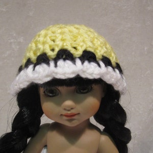 May include: A doll wearing a yellow, black, and white crocheted hat. The hat has a scalloped edge.