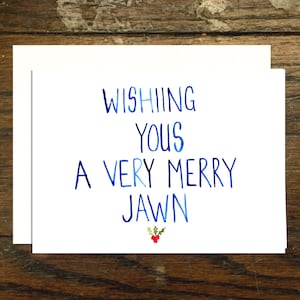 May include: A white greeting card with the text "Wishing Yous A Very Merry Jawn" in blue watercolor lettering. A small sprig of holly with red berries is at the bottom. The card is on a white envelope.