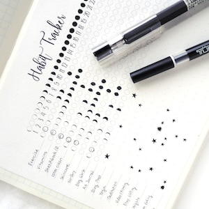 May include: A close-up of a habit tracker journal with a black pen. The journal page features a handwritten "Habit Tracker" title and a grid for tracking habits. The page is decorated with moon phases and star illustrations.