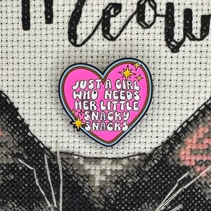 May include: A pink heart-shaped enamel pin with the text "JUST A GIRL WHO NEEDS HER LITTLE SNACKY SNACKS" in white lettering. The pin has a blue border and gold star accents. The pin is set against a white cross-stitch background with the word "meow" in black.