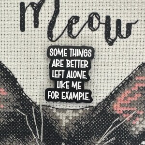 May include: Cross-stitch art with the word "Meow" stitched in black thread. A black and white pin with the text "SOME THINGS ARE BETTER LEFT ALONE. LIKE ME FOR EXAMPLE." is attached to the cross-stitch. The cat's face is partially visible.