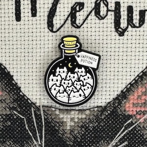 May include: A black and white enamel pin in the shape of a potion bottle. The bottle is filled with cat faces and has a label that reads "Happiness Potion". The bottle has a gold stopper.