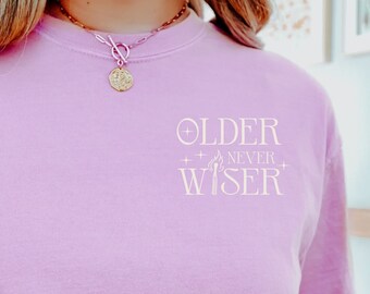 May include: A pink t-shirt with the text "OLDER NEVER WISER" printed on the front in white. The text is surrounded by small plus signs.