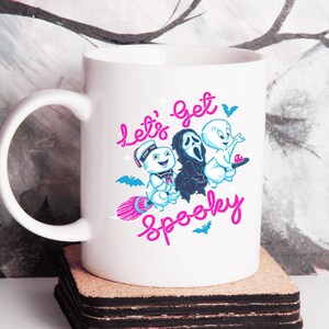 May include: White ceramic mug with a graphic of three ghosts. The ghosts are flying on a broomstick and the text "Let's get spooky" is written in pink.