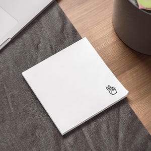 May include: A square white notepad with a pixelated peace sign in the corner. The notepad is on a dark grey surface next to a laptop and a potted plant on a wooden desk.