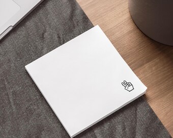 May include: A square white notepad with a pixelated peace sign in the corner. The notepad is on a dark gray surface next to a laptop and a potted plant on a wooden desk.