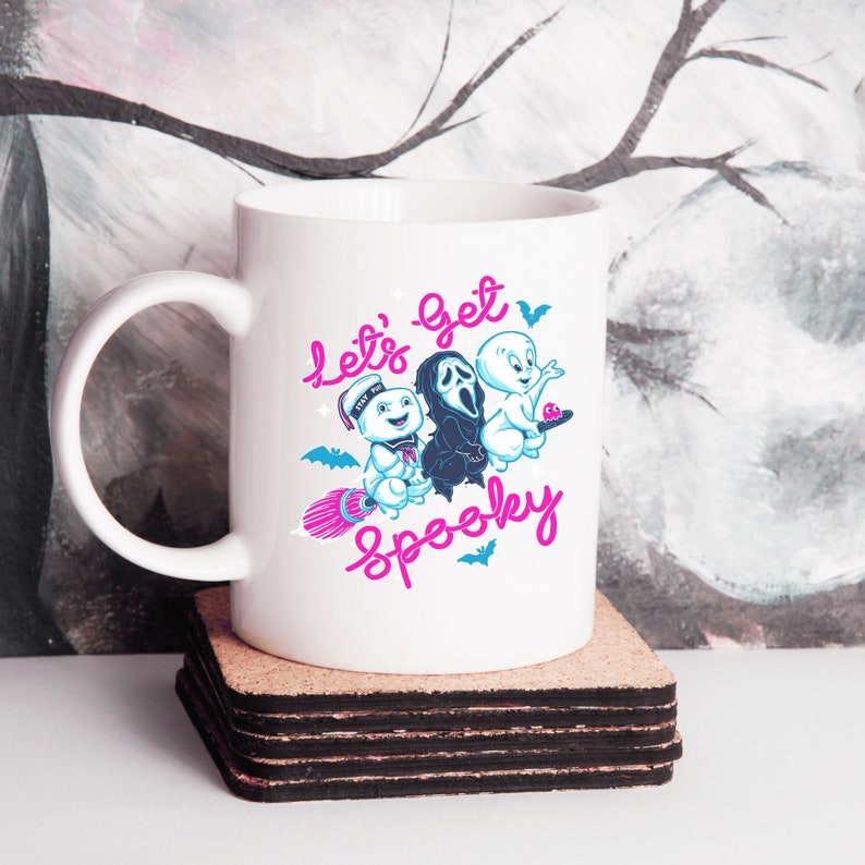 May include: White ceramic mug with a graphic of three ghosts. The ghosts are flying on a broomstick and the text "Let's get spooky" is written in pink.
