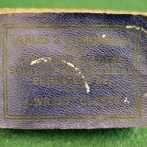 Puede incluir: Una pequeña etiqueta rectangular azul con letras doradas. El texto dice "TABLES & MEMORANDA FOR ENGINEERS, SOUTHERN ELECTRICAL ENGINEERING CO., J. WRIGHT, CLASS". La etiqueta tiene una superficie texturizada.