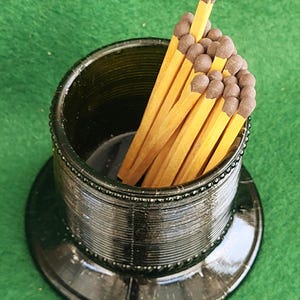 Puede incluir: Un portafósforos decorativo verde oscuro con base redonda y cuerpo cilíndrico. El portafósforos está lleno de varias cerillas de madera con puntas marrones. El portafósforos está sobre una superficie verde.