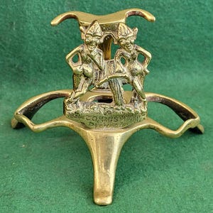 May include: A brass stand featuring two whimsical figures holding a mushroom. The stand has three legs and a decorative base.