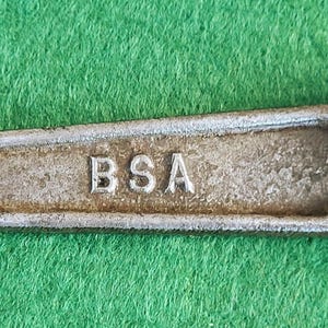 May include: A vintage metal wrench with a hexagonal opening on each end. The wrench is a dull silver color with the letters "BSA" stamped in the center. The wrench is lying on a green surface.
