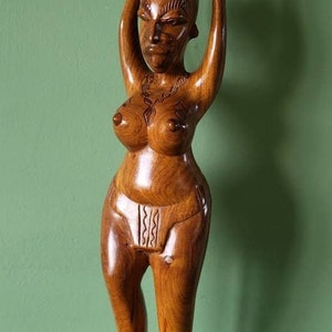 May include: A wooden sculpture of a woman carrying a pot on her head. The figure is carved from light brown wood, with detailed facial and body carvings. The sculpture has a smooth, polished finish and stands on a round base. The sculpture is approximately 30 cm tall.