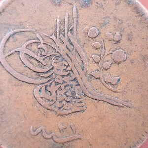 May include: Close-up of a copper coin with Arabic script and a floral design. The coin is worn and has a brown patina.