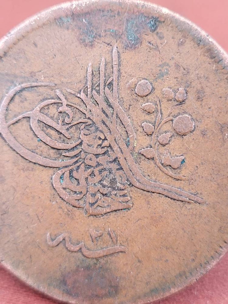 May include: Close-up of a copper coin with Arabic script and a floral design. The coin is worn and has a brown patina.