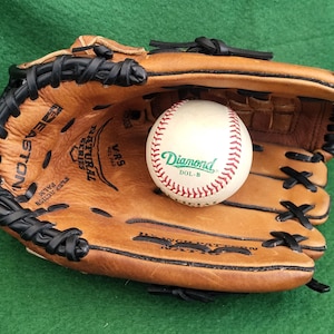 May include: A baseball glove and baseball on a green surface. The baseball is white with red stitching and the word "Diamond" in green. The glove is brown leather with black stitching and accents. The glove is open, with the baseball resting inside.