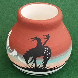 Può includere: Un piccolo vaso rotondo in ceramica color terracotta con un'ampia apertura. Il vaso presenta una silhouette nera di un uccello con macchie bianche, su uno sfondo simile a un tramonto con strisce arancioni, blu e bianche. La base ha strisce bianche e nere.