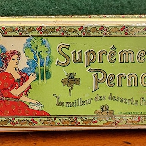 Puede incluir: Caja de hojalata vintage con fondo verde y letras doradas. La caja presenta a una mujer con un vestido rojo y el texto "Suprême Pernod" y "Le meilleur des desserts fins..."