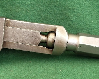 Puede incluir: Una herramienta metálica antigua con una cabeza triangular y una mordaza ajustable. La herramienta tiene un cuerpo cilíndrico y un mango hexagonal. El metal tiene un aspecto desgastado y envejecido, lo que sugiere que es una antigüedad o un artículo muy usado.