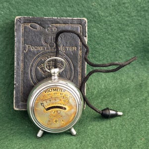 May include: A vintage silver-toned pocket voltmeter with a round face, yellowed dial, and black needle. The device features the text VOLTMETER, DIRECT CURRENT, J.T., FOREIGN, B 120, and A 12. It includes a black cord and rests before its black case.