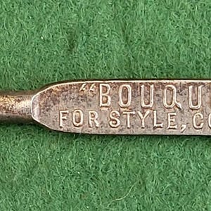 Peut inclure: Un chausse-pied argenté vintage avec une poignée en forme de boucle ovale. La poignée porte l'inscription "BOUQUET" BOOTS FOR STYLE, COMFORT & WEAR". Le chausse-pied est posé sur un fond vert.