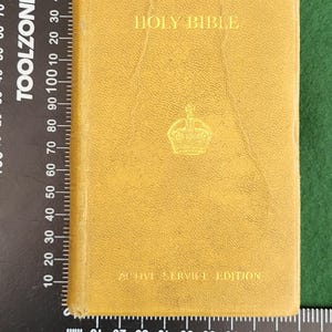 May include: A mustard-colored Holy Bible with gold lettering and a crown emblem. The text "HOLY BIBLE" is at the top, and "ACTIVE SERVICE EDITION" is at the bottom. The book is rectangular and appears to be vintage.