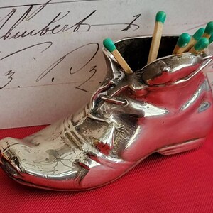 Puede incluir: Porta cerillas de plata con forma de zapato con cerillas de punta verde en su interior.