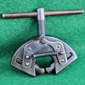 May include: A vintage metal tool with a T-shaped handle and a spring-loaded clamping mechanism. The tool is made of dark metal with a worn appearance, suggesting age and use. The background is a solid green.
