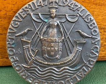May include: A bronze medal commemorating the 1947 Lisbon Games. The medal features a woman holding a shield with the Portuguese coat of arms, surrounded by a wreath of laurel leaves. The inscription "PORTUGUESES 1947 LISBOA 1947" is visible around the edge of the medal.